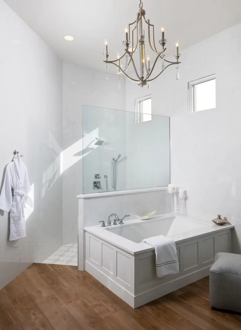 Elegant soaking tub with panel surround, chandelier above, wood floor, shower behind glass